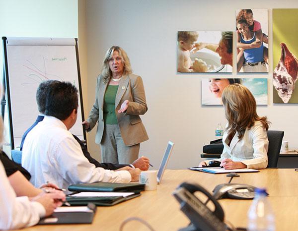 An adult woman presenting information from a chart to people gathered around a conference table.